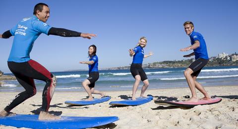 兩名年輕女孩與一名年輕男孩正在邦迪海灘，向「Let's Go Surfing」課程的導師學習滑浪